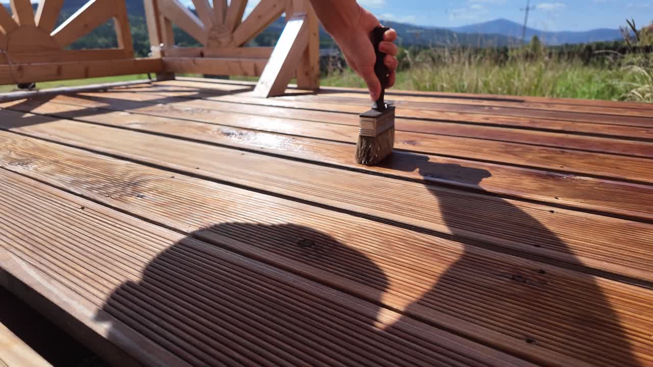 Hand oils varnishing impregnating wooden terrace boards with brush. Decorative railing, serene sky, clouds, and mountain horizon evoke home improvement and outdoor maintenance themes - 4K
