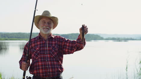 senior fisherman in a hat posing for the camera and smiling with a fish on a rod on the shore of a lake in the morning