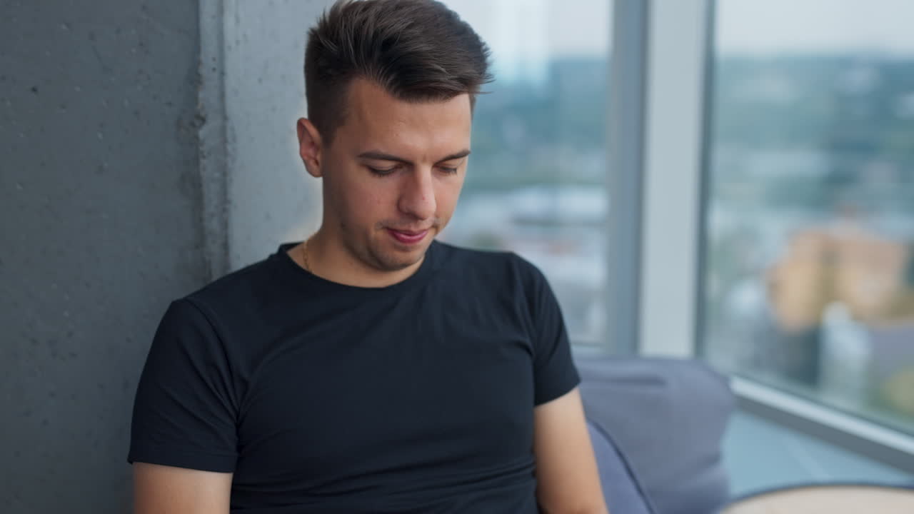 Dark-haired young man focused on his work at laptop. Male sitting near the panoramic window in a comfortable chair. Blurred backdrop.