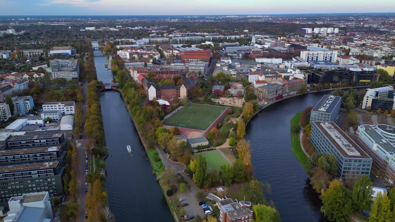 Berlin cityscape featuring a river, urban buildings, and a soccer field during autumn. Smooth aerial view flight drone shot from above
