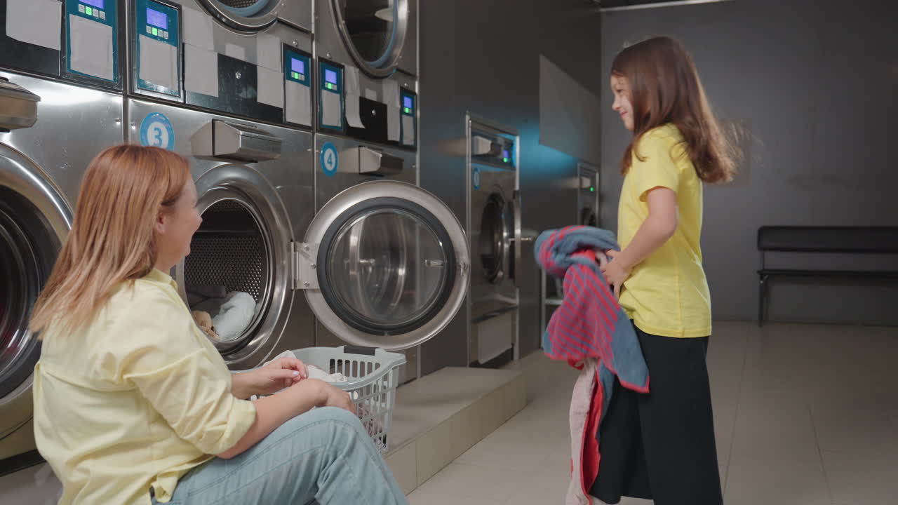 House wife tosses cloth to playful child, child throws fabric into washing machine inside laundromat, mother sits near open door with basket of towels, sharing teamwork, hygiene routine, family chore