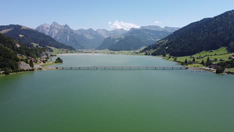 Toma-Aérea-De-Autos-Moviéndose-En-Un-Puente-Sobre-El-Lago-Sihlsee-Con-Una-Cordillera-Detrás