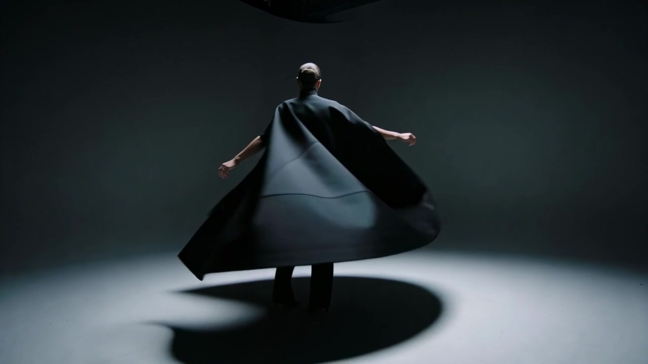 Levantando los brazos, modelo girando bajo un foco en un estudio, con una capa negra escultural creando un halo oscuro.