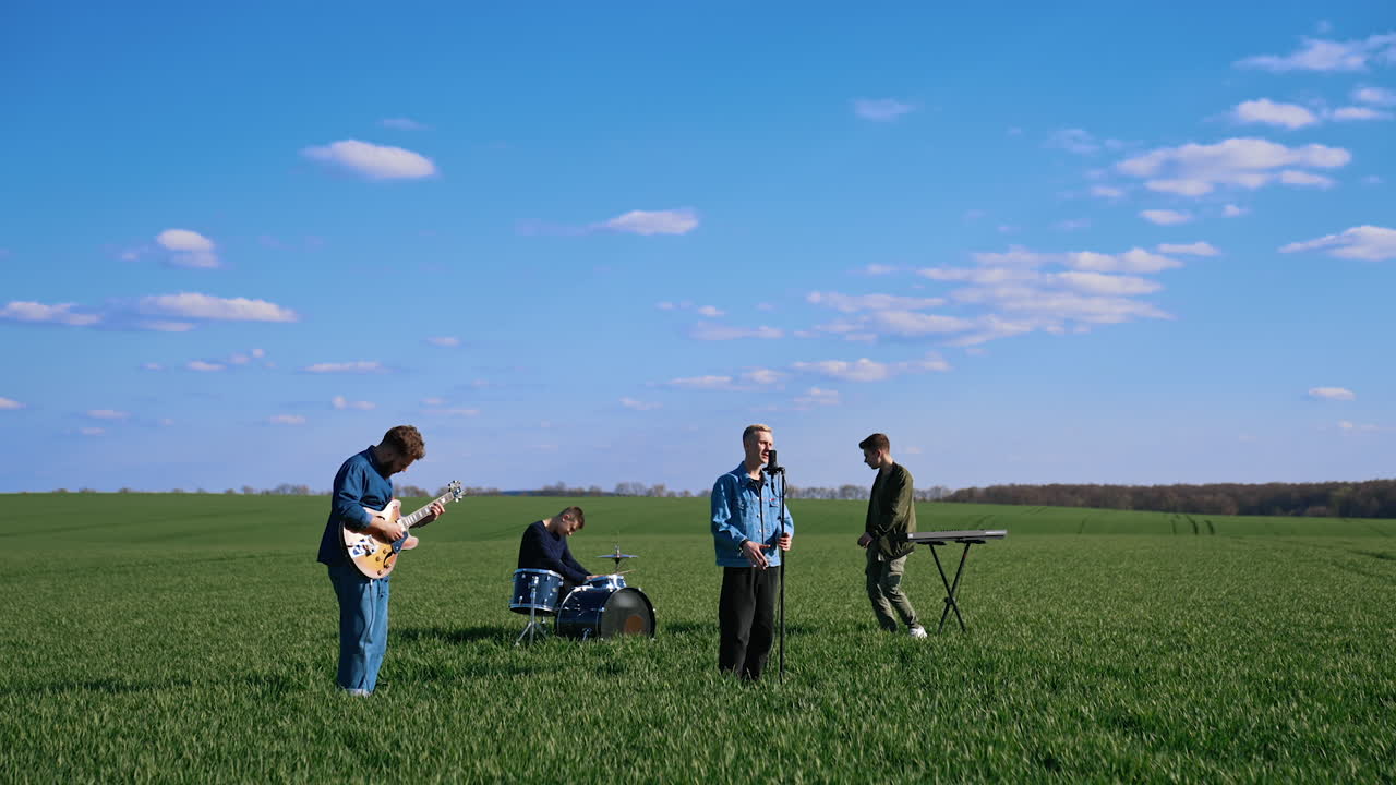 Outdoor concert of young musicians. Group of friends playing the guitar, drums and synthesizer performing music on green field under blue sky.