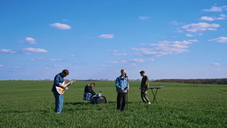 outdoor concert of young musicians. group of friends playing the guitar, drums and synthesizer performing music on green field under blue sky.