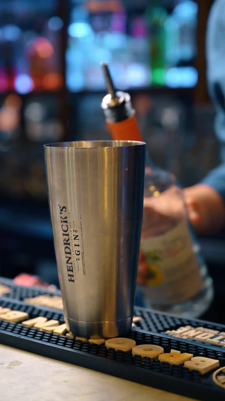 New York, USA, 15 aprile 2026: Shaker da cocktail professionale in metallo su tappetino da bar in gomma nera. Bicchiere shaker da cocktail in acciaio appoggiato su un tappetino da bar testurizzato con sfondo sfocato di bottiglie all'interno di un bar