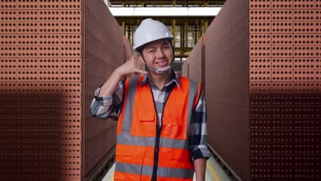 asian male engineer with safety helmet smiling to camera and making call me gesture while standing with red brick packed in stacks are stored