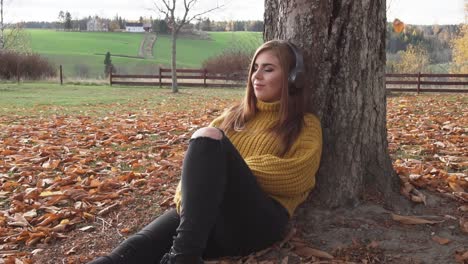 adult girl is sitting under the tree and listening to audio books or music in autumn scenery and relaxing