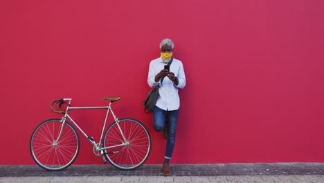 hombre mayor afroamericano que usa una máscara facial con bicicleta usando un teléfono inteligente mientras está de pie contra p