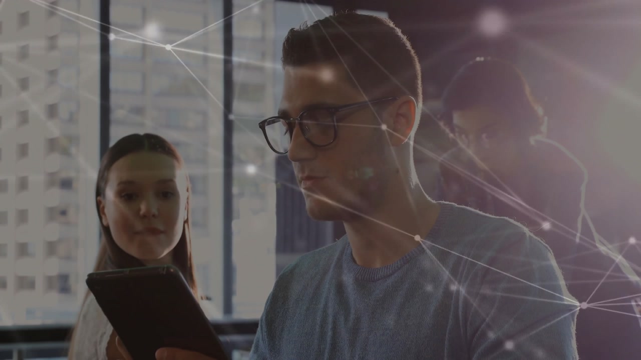 Male colleague scrolling tablet by window while female colleague joins, reviewing tech data overlay