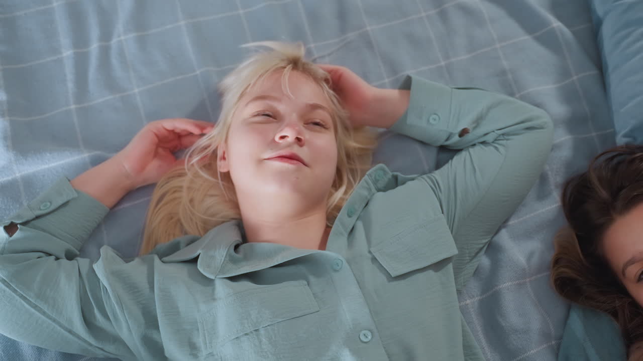 Blonde girl in loose mint sleepwear lies on blue bed, smiling and touching hair joyfully while subtly shaking body, another person lies beside her as they enjoy relaxed