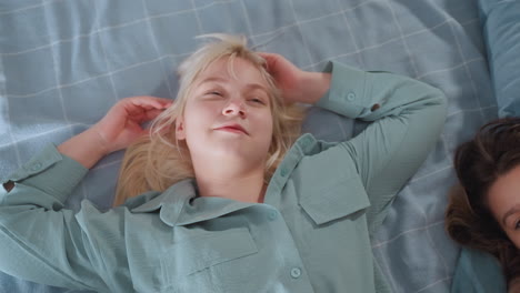 blonde girl in loose mint sleepwear lies on blue bed, smiling and touching hair joyfully while subtly shaking body, another person lies beside her as they enjoy relaxed