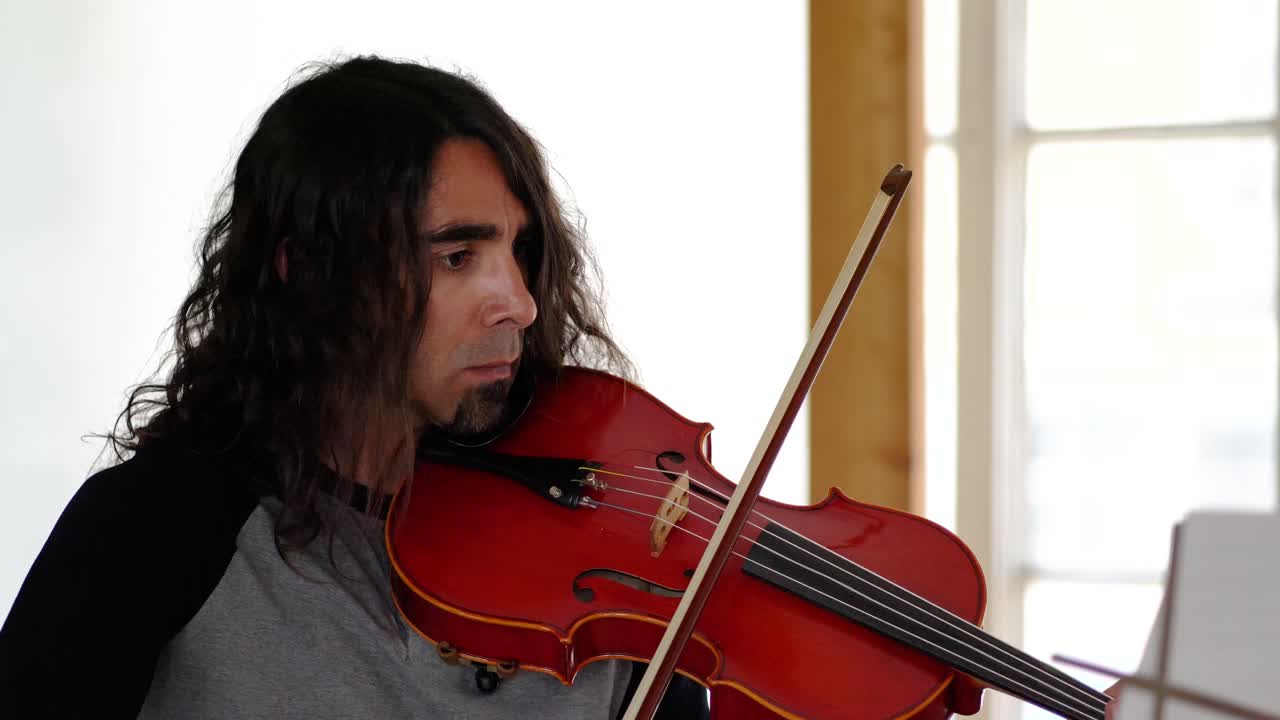 cerca de un músico latino con cabello largo y castaño interpretando tranquilamente una canción en una hermosa viola roja en un estudio blanco.