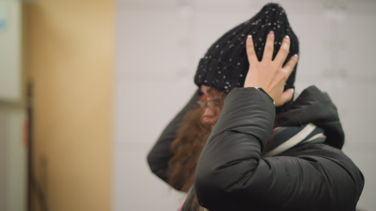 Damsel wearing black jacket and striped scarf adjusts curly hair while tilting head down, hand lifting strands with casual movement, as she wears her head cap smartwatch on wrist visible