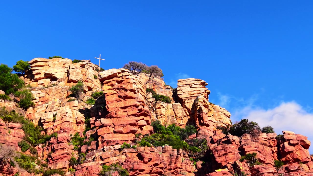 Formazione rocciosa di arenaria rossa con croce di legno sulla cima, Spagna. Vista dal basso di una parete rocciosa arancione stratificata con rada vegetazione verde contro un cielo blu intenso e nuvole leggere all'orizzonte.
