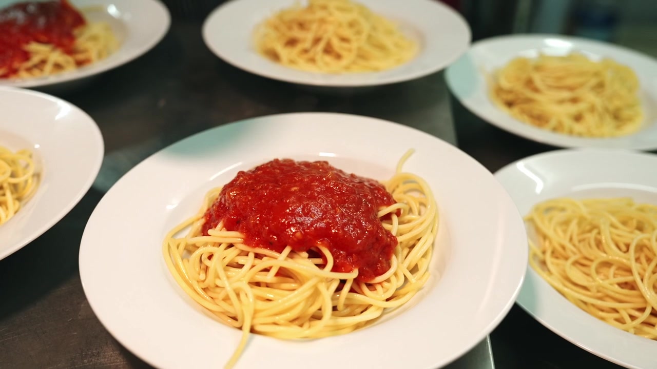 Carrellata Macro di uno Chef che Usa un Mestolo di Metallo per Versare una Densa Salsa Marinara di Pomodoro Rosso Vivo su Mucchi di Spaghetti Cotti su Piatti Bianchi Allineati su un Bancone della Cucina