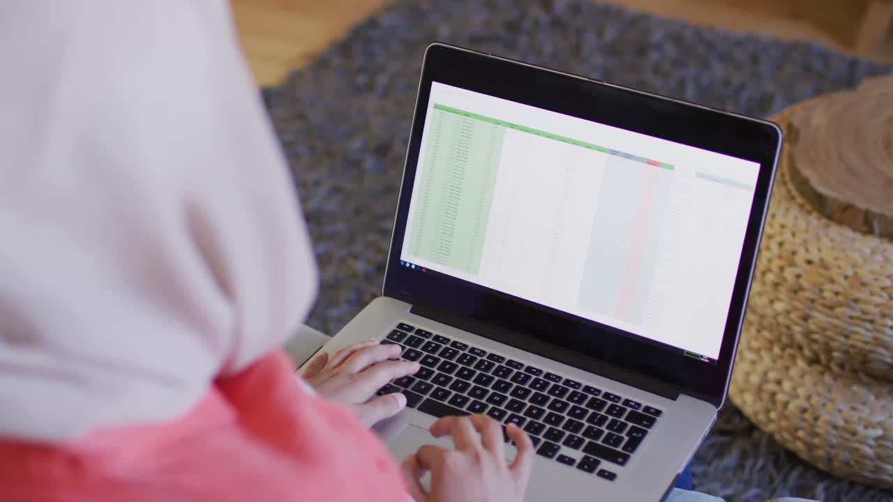 Over shoulder video of biracial woman in hijab at home working on laptop, spreadsheet on screen