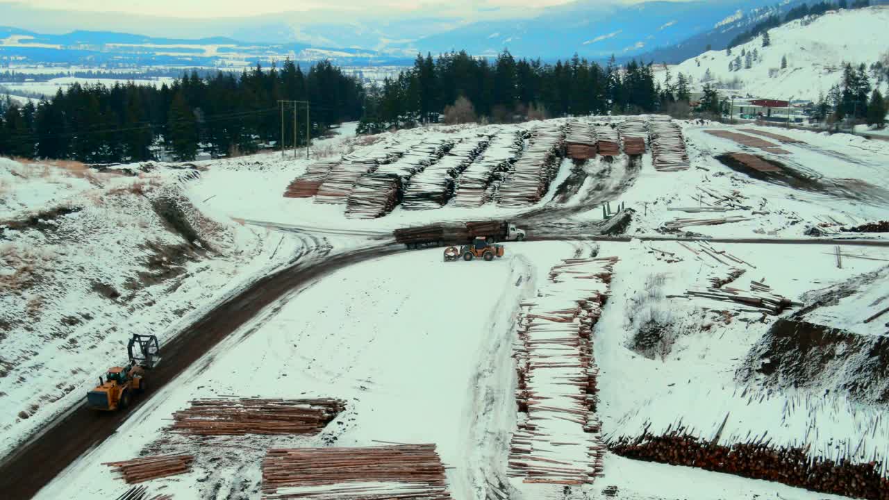 Aerial follow shot of wheel loader with grapple bucket maneuvering towards log truck to unload logs at Canadian sawmill in winter