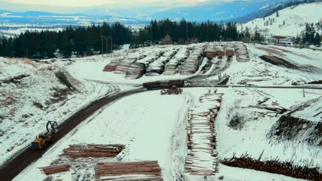 Luftaufnahme-Eines-Radladers-Mit-Greifschaufel,-Der-In-Richtung-Eines-Holzlastwagens-Manövriert,-Um-Im-Winter-Holz-Im-Kanadischen-Sägewerk-Zu-Entladen