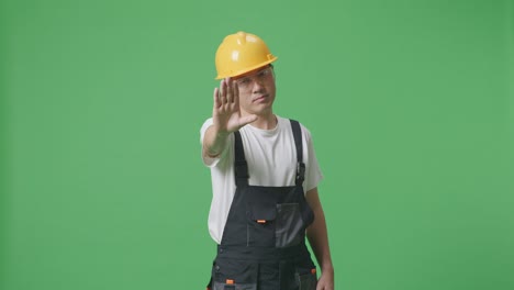 hombre asiático trabajador con gafas de protección y casco de seguridad mirando a la cámara y desaprobando sin señal de mano mientras está de pie en el estudio de fondo de la pantalla verde