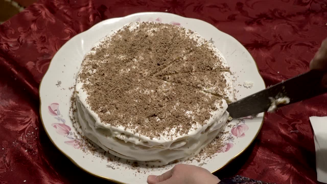 una mujer corta en pedazos iguales un pastel blanco festivo decorado con chocolate rallado en la parte superior. el pastel vale la pena en un plato blanco y manteles rojos. primer plano. la vista desde lo alto. 4k.