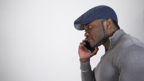 serious afro american guy talking the phone. young black man in blue stylish cap and gray sweater having a conversation on a phone isolated on white. profile view.