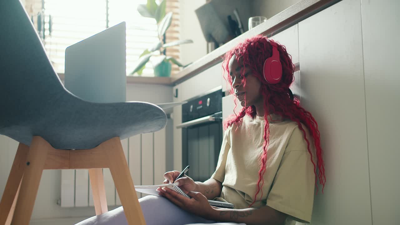 African American girl with red curly hair sitting on kitchen floor with laptop and making notes