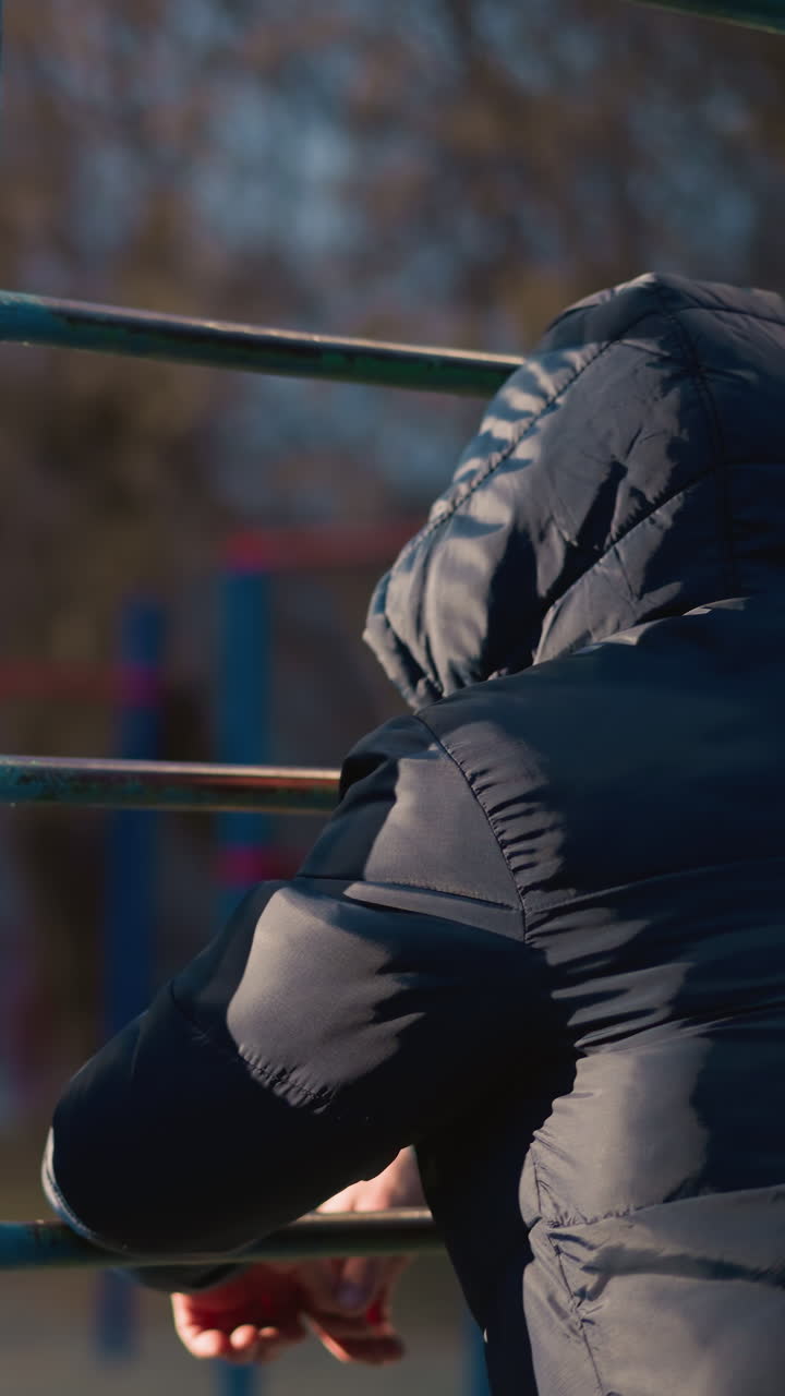Close-up of two people resting their hands on an iron bar, both wearing a hooded jacket and the other in the background blurred, with a blurred building in the distance