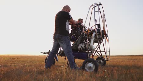 motor paraglider stands in the field at sunset with a wooden propeller, two pilots warm up the engine before the flight