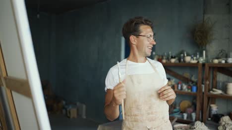 Man-in-glasses-paints-a-picture-and-singing,-having-fun.-A-talented-art-worker,-designer-in-his-studio