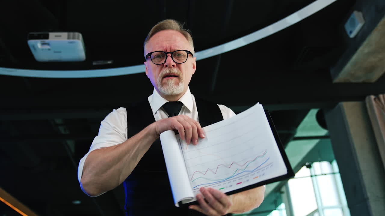 Senior male wearing white shirt and black vest holding the documents. Businessman turns the pages displaying charts. Low angle view.