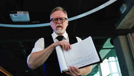 senior male wearing white shirt and black vest holding the documents. businessman turns the pages displaying charts. low angle view.