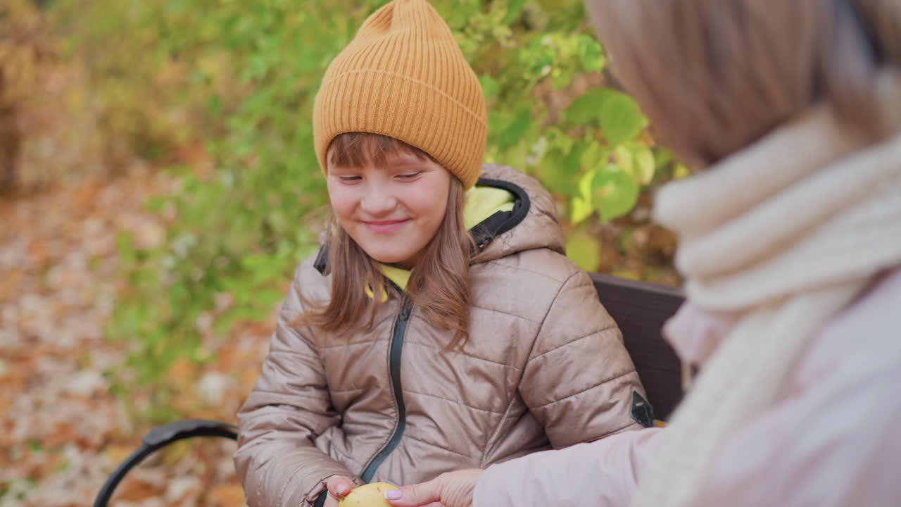 Young girl in brown jacket and orange beanie sits quietly on park bench, gazing down with soft expression as caring mother in scarf gently hands her fruit during peaceful autumn day