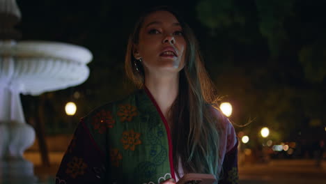 mujer esperando reunión de teléfono inteligente de pie cerca de la fuente por la noche de cerca.