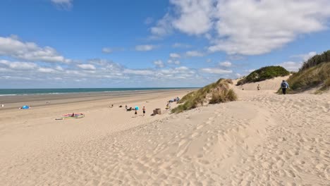 wide shot of a sandy beach with scattered people relaxing under clear blue skies, gentle dunes, and calm sea. natural daylight, static camera