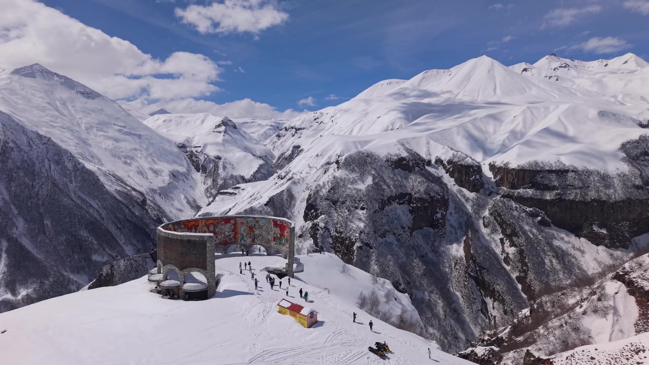 Filmato drone che orbita attorno al Monumento dell'Amicizia Georgia-Russia a Gudauri, rivelando catene montuose innevate e un drammatico paesaggio invernale sullo sfondo.