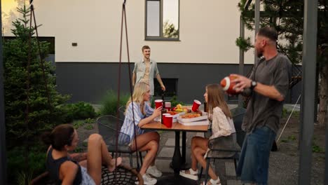 Happy-blond-guy-with-a-mustache-throws-an-American-football-ball-to-his-friend-while-relaxing-at-a-table-in-the-backyard-of-a-house