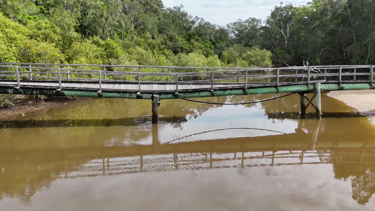 Camera smoothly moves across rustic footbridge above brown river, lush green bushland, daylight