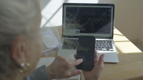 elderly-senior-woman-holding-smartphone-and-laptop-with-stock-market-investment-analysis-trading-online-in-slow-motion