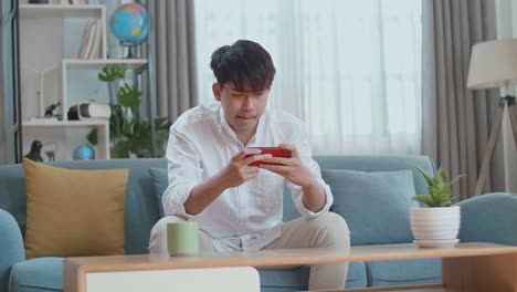 Asian Man Being Upset Losing Game On Smartphone While Sitting On Sofa In The Living Room