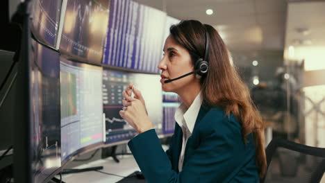 una mujer de negocios con traje y auriculares está mirando una pantalla de computadora con gráficos y gráficos, y hablando en un micrófono.