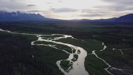 Impressive-flight-above-zigzag-Yukon-Dezadeash-river-and-dark-green-valley-with-rocky-rough-Saint-Elias-mountain-range-in-background-with-bright-white-sun-at-sunset,-Canada,-overhead-drone-approach