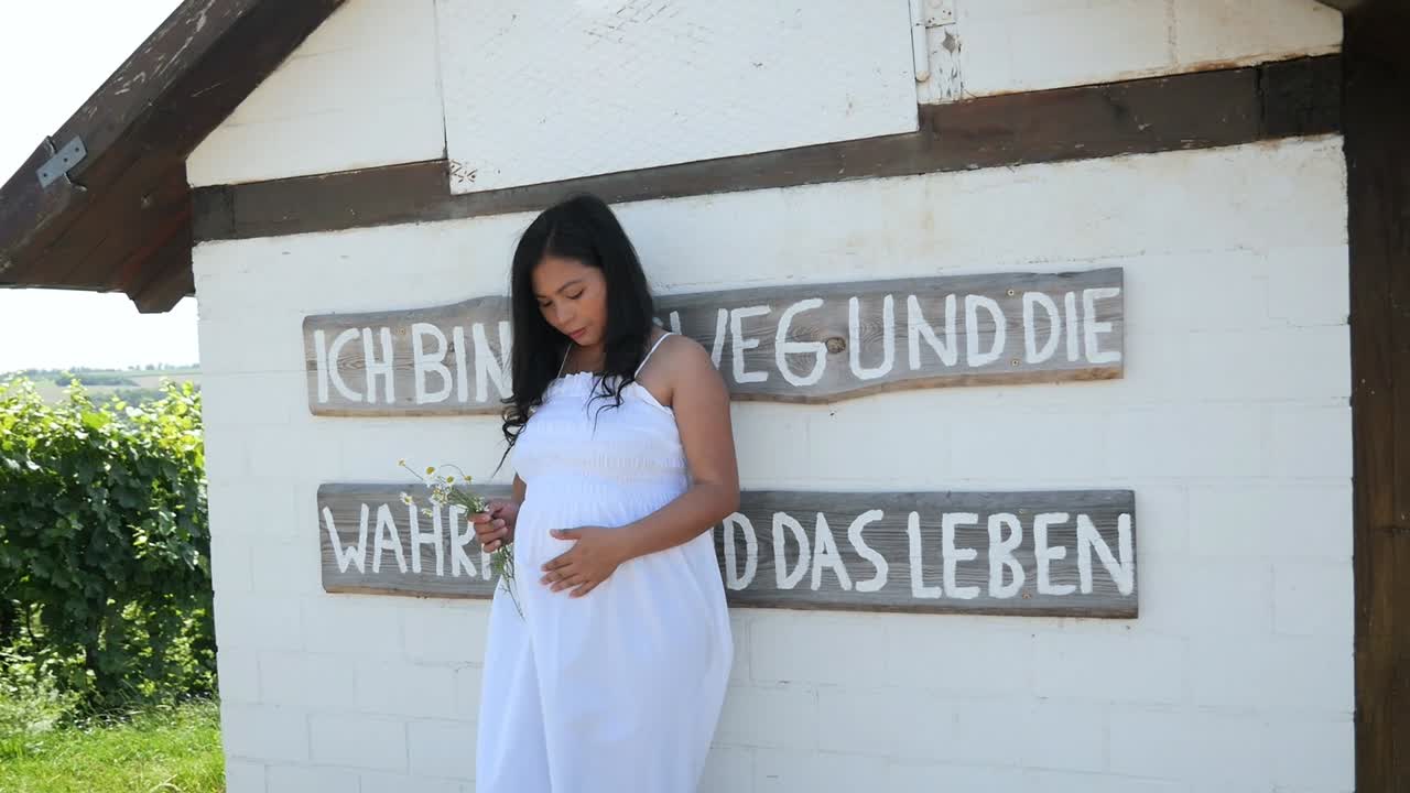 Ung gravid kvinde i en hvid sommerkjole, der læner sig op ad en mur med blomster i hænderne til et graviditetsfotoshoot.