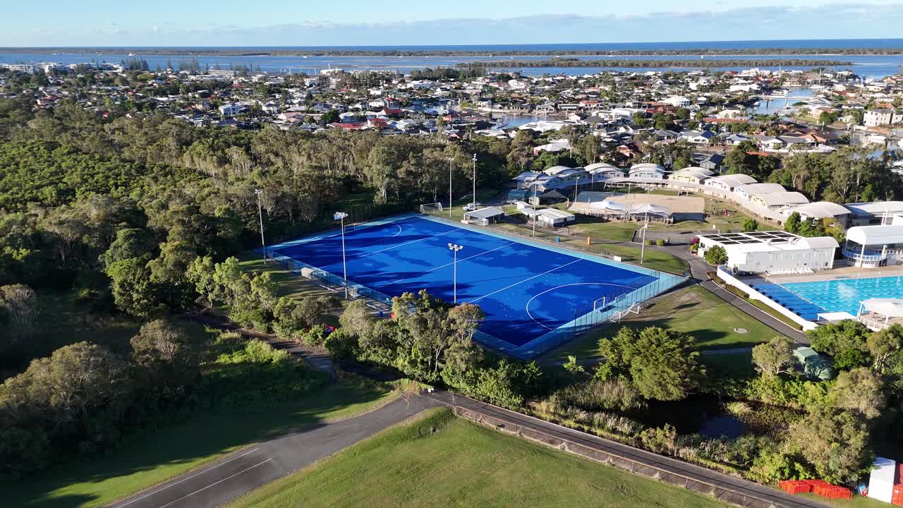 Drone footage captures a vibrant sports park with blue fields surrounded by greenery and urban landscape under clear skies