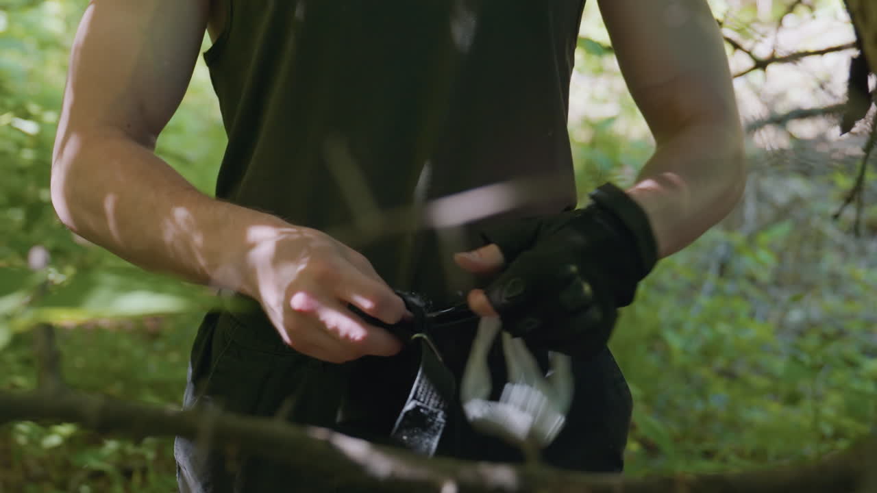 soldier wearing fingerless glove grips cloth headband with bare forearm visible while adjusting strap under dappled forest light near tree bark conveying rugged survival preparation and focused