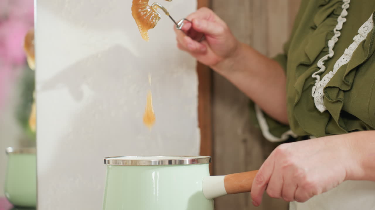 Partial view of white chef stirring soup in light green pot using metal spoon while wearing green ruffled dress and apron in rustic kitchen with wooden elements and soft natural lighting