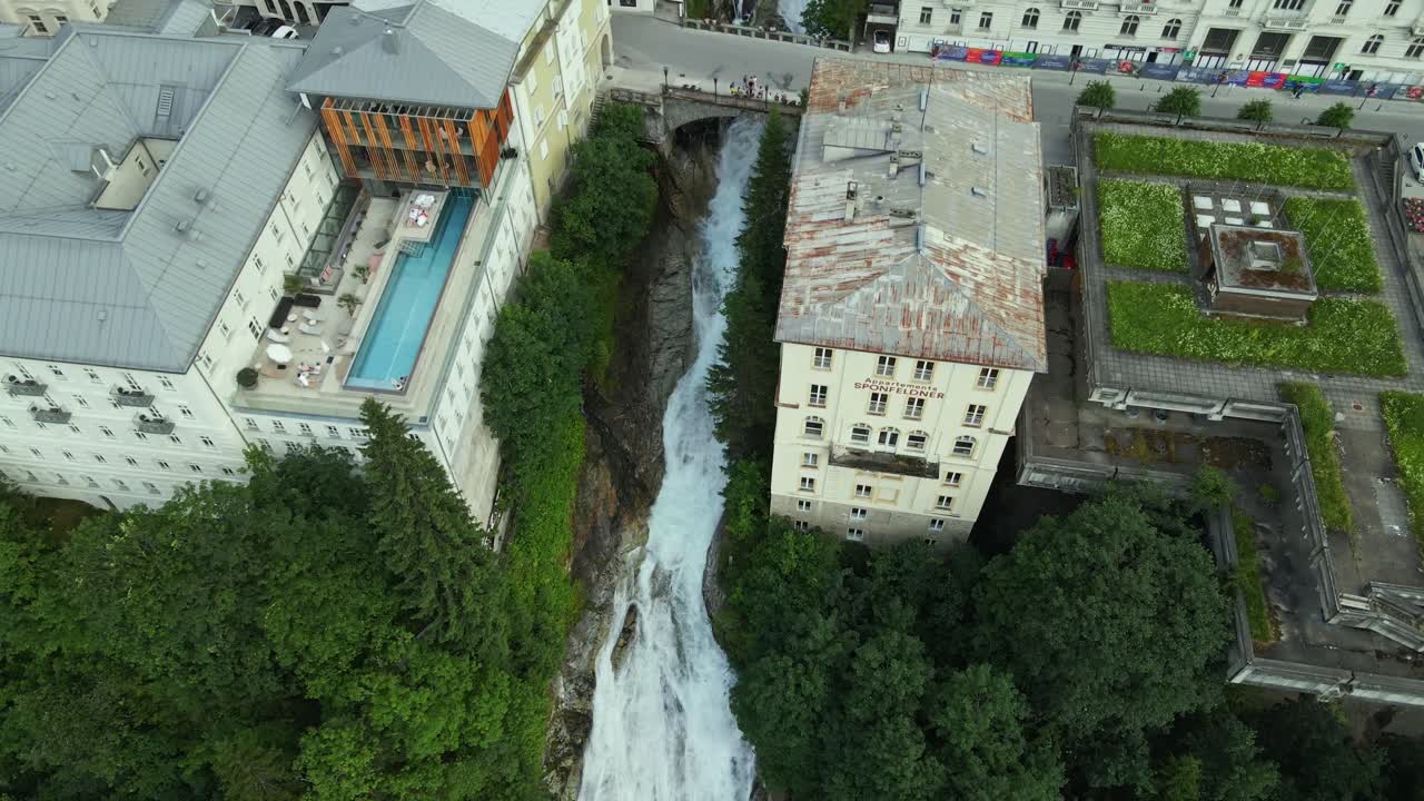 A waterfall flows through the center of a town, surrounded by historic buildings and modern structures. The contrast between natural force and architectural beauty creates a unique urban landscape.