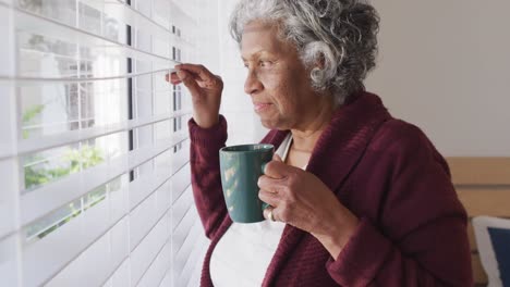 Happy-senior-african-american-woman-looking-through-window-and-drinking-coffee