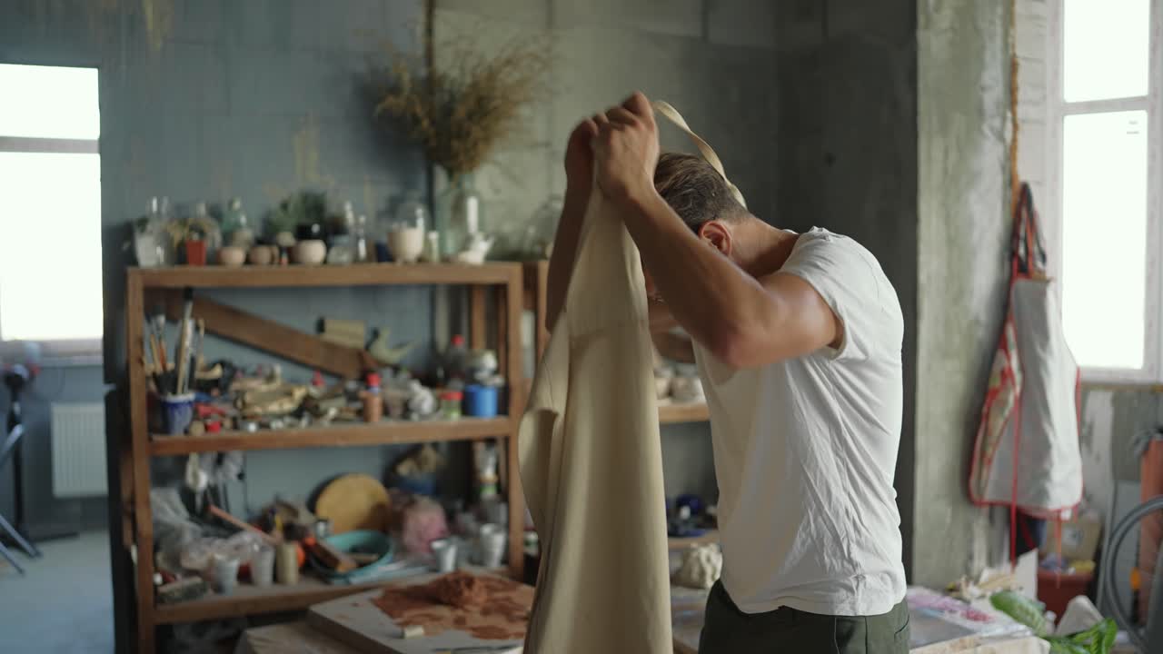 A happy successful potter in a studio studio finishes his working day in the background of a shelf with his products, takes off