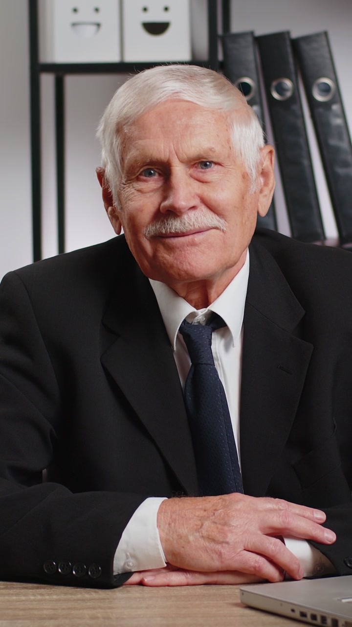 Retrato de un empresario de alto rango en traje sentado en una mesa con una computadora portátil sonriendo a la cámara en la oficina de su casa